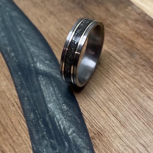 May include: A silver ring with a dark, textured inlay and two thin silver bands. The ring is set against a wooden surface with a dark blue, glossy accent.