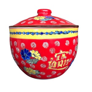 Op de afbeelding: Rode keramische pot met een deksel, versierd met een bloemenpatroon en Chinese karakters. De pot heeft een geel en blauw bloemenpatroon en een gele en blauwe rand.