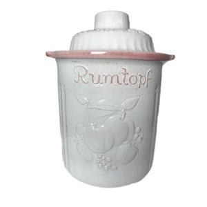 Könnte beinhalten: Weiße Keramikdose mit rosafarbenem Deckel und rosafarbenem Rand. Die Dose hat ein Reliefdesign mit Früchten und dem Wort "Rumtopf" auf der Vorderseite.