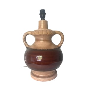 Puede incluir: Una lámpara de mesa de cerámica marrón y beige con dos asas. La lámpara tiene un esmalte marrón en la mitad inferior y un esmalte beige en la mitad superior. La lámpara tiene una pantalla negra simple.