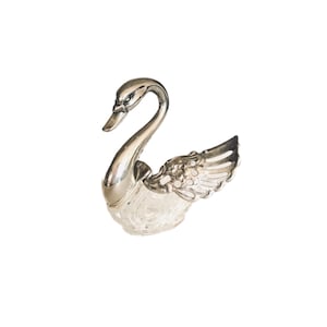 May include: A silver swan figurine with a clear, textured glass base. The swan has detailed wings and a curved neck, creating an elegant and decorative piece. The figurine is set against a white background.