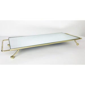 May include: A rectangular mirrored tray with a gold-coloured metal frame and legs. The tray has a handle on one end and is supported by four small ball-shaped feet. The mirrored surface reflects light, creating a bright and elegant look.