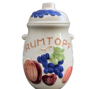 May include: White ceramic jar with a lid, decorated with a grapevine design and the word "RUMTOPF" in orange lettering. The jar has two handles and is decorated with fruit and grapes.
