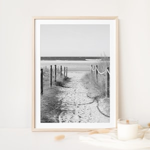 May include: Black and white framed print of a sandy path leading to the ocean. The path is bordered by wooden posts and rope, with tall grass on either side. The sky is clear and the ocean is visible in the distance.