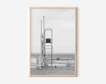 Puede incluir: Fotografía en blanco y negro de una torre de salvavidas en un muelle, con el océano y el cielo de fondo. La torre es de metal y tiene una escalera que conduce a una pequeña plataforma. La imagen está enmarcada en un marco de madera clara.