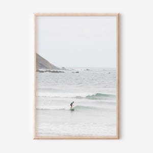 Op de afbeelding: Een ingelijste print met een surfer op een golf in de oceaan. De afbeelding heeft een gedempt kleurenpalet met een lichtblauwe lucht en water. Op de achtergrond is een kustlandschap te zien.
