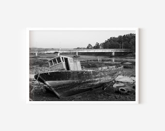 May include: Black and white photograph of a weathered wooden boat beached on muddy ground. A bridge spans the background, with trees lining the horizon. The boat's cabin is visible, and the scene evokes a sense of age and abandonment.