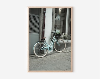 Op de afbeelding: Een ingelijste print van een lichtblauwe vintage fiets, geparkeerd op een geplaveide straat. De fiets heeft een bruin zadel, een zwarte mand gevuld met bloemen en crèmekleurige banden. De achtergrond toont een gebouw met een grijze deur.