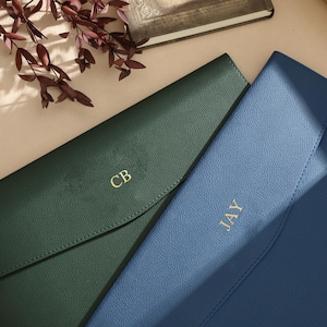 May include: Two leather document holders, one in forest green and the other in blue. The green holder has the initials "CB" in gold, while the blue one has "JAY". Both are closed, with a flap closure. The holders are on a light-coloured surface.