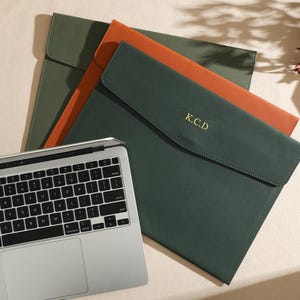 Può includere: Tre custodie per laptop in pelle verde e arancione, con una tastiera argentata in primo piano. La custodia verde ha le iniziali "K.C.D" in oro. Le custodie sono chiuse con un lembo.