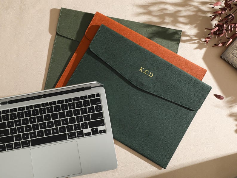 May include: Three leather laptop sleeves in green and orange, with a silver laptop keyboard in the foreground. The green sleeve has the initials "K.C.D" in gold. The sleeves are closed with a flap.