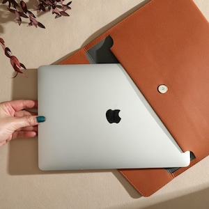 May include: A silver laptop with a black Apple logo is partially inserted into a tan leather laptop sleeve. The sleeve is open, revealing a grey interior and a magnetic closure. A hand with teal nail polish is holding the laptop.