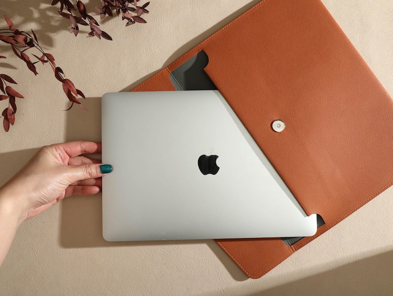 May include: A silver laptop with a black Apple logo is partially inserted into a tan leather laptop sleeve. The sleeve is open, revealing a grey interior and a magnetic closure. A hand with teal nail polish is holding the laptop.
