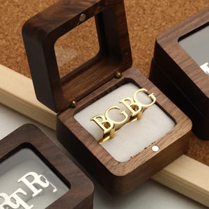 Może przedstawiać: Złoty pierścionek z napisem "BGBG" w otwartym drewnianym pudełku na biżuterię z przezroczystą pokrywką. Pierścionek spoczywa na białej poduszce. W tle widoczne są inne zamknięte pudełka z literami.