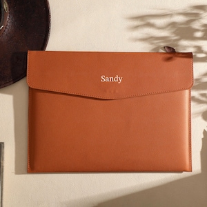 May include: A leather laptop sleeve in a warm orange hue, with a flap closure. The word "Sandy" is subtly printed on the front. A brown hat and a pen are visible in the background, suggesting a workspace or desk setting.