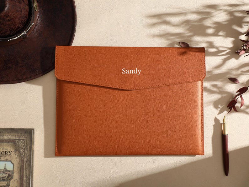 May include: An orange leather laptop sleeve with a flap closure, bearing the name "Sandy". A brown hat and a pen are in the background. The scene suggests a workspace or desk setting, with a focus on the sleeve.