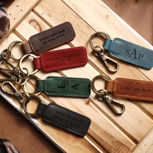 May include: Several leather keyrings in shades of brown, red, green, blue, and black. Each keyring has a metal clasp and ring, with engraved messages and initials. The keyrings are displayed on a wooden surface.
