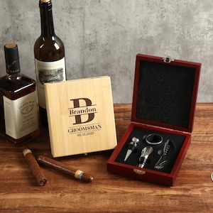 Puede incluir: Una caja de madera grabada con "Brandon GROOMSMAN 08.18.2025" junto a una botella de licor, una botella de vino, dos puros, una copa de vino y una caja de madera roja que contiene herramientas para vino.