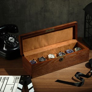 Może przedstawiać: Drewniana szkatułka na zegarki z otwieranym wiekiem, ukazująca sześć miejsc na zegarki. Szkatułka wykonana jest z ciemnego drewna z jasnym wzorem słojów. Szkatułka stoi na ciemnym drewnianym blacie.