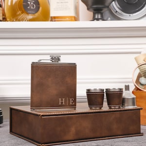Puede incluir: Una petaca envuelta en cuero marrón con las iniciales "H|E" grabadas, en una caja de cuero marrón con dos vasos de chupito y un embudo.