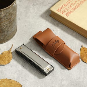 Op de afbeelding: Een zilveren mondharmonica met een bruine leren hoes. De hoes heeft het woord "Haley" erop gegraveerd.