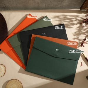 May include: A selection of document holders in various colours: orange, green, black, brown, and dark green. Each has a flap closure, with initials on some. A brown leather hat and wood slices are also present.