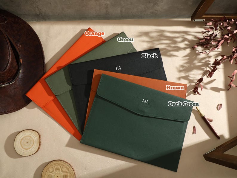 May include: A selection of document holders in various colours: orange, green, black, brown, and dark green. Each has a flap closure, with initials on some. A brown leather hat and wood slices are also present.
