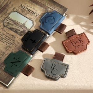 Puede incluir: Un conjunto de marcadores de cuero personalizados en varios colores, incluyendo azul, negro, verde, gris y marrón. Cada marcador presenta una inicial, nombre o fecha únicos, sujetos a una banda elástica marrón. Los marcadores se muestran sobre un libro de estilo vintage.
