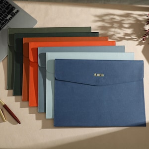 Peut inclure: Plusieurs pochettes en cuir de différentes couleurs, dont vert, orange et bleu. La pochette bleue au premier plan porte le nom "Anna" en doré. Un ordinateur portable et un stylo sont également visibles.