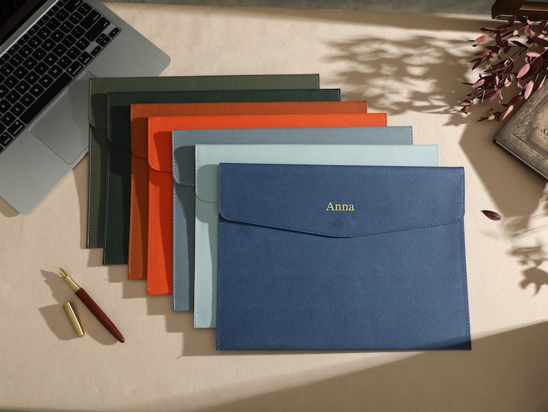 May include: A selection of leather document holders in shades of green, orange, and blue. The front blue holder is embossed with the name "Anna" in gold. A laptop and pen are also visible.