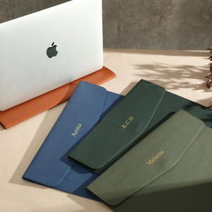 May include: A white laptop with a black Apple logo sits on a brown leather laptop sleeve. Three personalised leather document holders in blue, green, and olive green are displayed. Each holder has a name or initials in gold lettering.