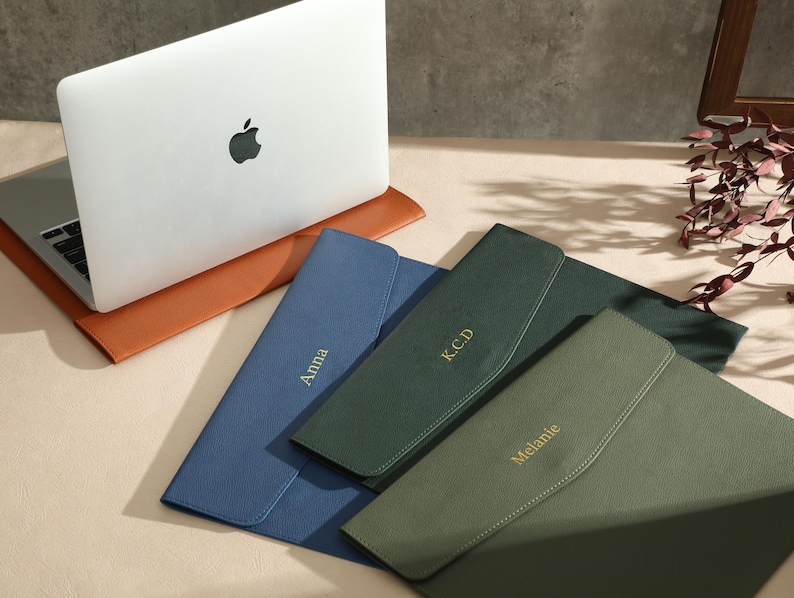 May include: A white laptop with a black Apple logo sits on a brown leather laptop sleeve. Three personalised leather document holders in blue, green, and olive green are displayed. Each holder has a name or initials in gold lettering.