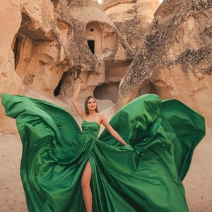 May include: A woman in a flowing emerald green strapless gown poses in a canyon. The dress has a high slit and the fabric is billowing in the wind. The background features unique rock formations in shades of tan and brown.