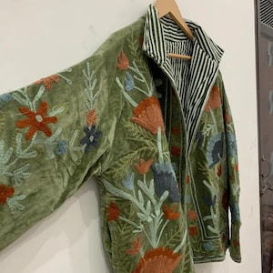 Pu&ograve; includere: Giacca ricamata verde oliva con disegni floreali arancioni, blu e verdi. La giacca presenta una fodera a righe e un colletto. Il ricamo &egrave; dettagliato e la giacca &egrave; appesa a una gruccia di legno.