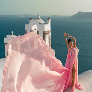 Può includere: Un lungo abito rosa chiaro con uno spacco alto e un lungo strascico fluente. L'abito è indossato su un tetto bianco con una chiesa sullo sfondo, con vista sul mare. Il tessuto ondeggia nel vento.