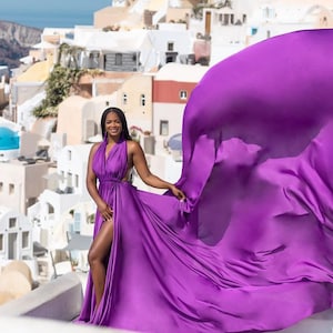 May include: A woman in a vibrant purple halter dress with a flowing skirt, posing against a backdrop of white buildings and blue domes. The dress features a high slit, and the fabric billows in the wind, creating a dramatic effect. The background includes a view of the sea.