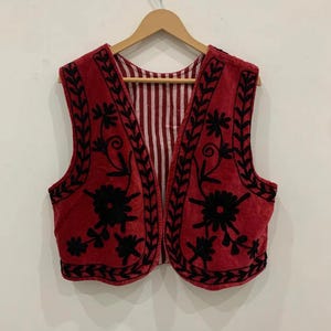 Puede incluir: Un chaleco rojo con bordados florales negros. El chaleco tiene cuello en V y es sin mangas. El forro interior presenta un patrón de rayas blancas y rojas. El chaleco está colgado en una percha de madera.