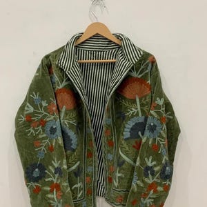 Könnte beinhalten: Olivgrüne Jacke mit Blumenstickerei in Orange, Blau und Rot. Die Jacke hat einen gestreiften Kragen und ein gestreiftes Innenfutter. Die Jacke hängt an einem Holzbügel.