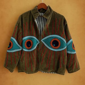 Puede incluir: Chaqueta verde oliva con grandes diseños de ojos. La chaqueta presenta apliques de ojos turquesa y naranja con líneas rojas radiantes. Tiene cuello y forro a rayas blancas y negras. Cuelga de una percha de madera.