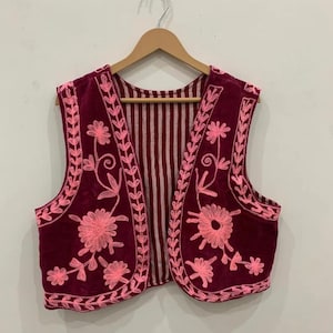 Pode incluir: Um colete de veludo bordô com bordados florais rosa. O colete tem forro listrado e é exibido em um cabide de madeira. O bordado apresenta padrões florais detalhados nas bordas e painéis frontais.