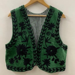 May include: Green vest with black floral embroidery. The vest has a black and white striped lining and is hanging on a wooden hanger. The vest is sleeveless and has a V-neck.