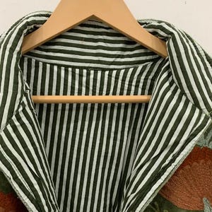 Pu&ograve; includere: Primo piano di una giacca verde con interno e colletto a righe. La giacca &egrave; appesa a una gruccia di legno. La giacca ha un motivo floreale ricamato nei toni dell'arancione e del verde.