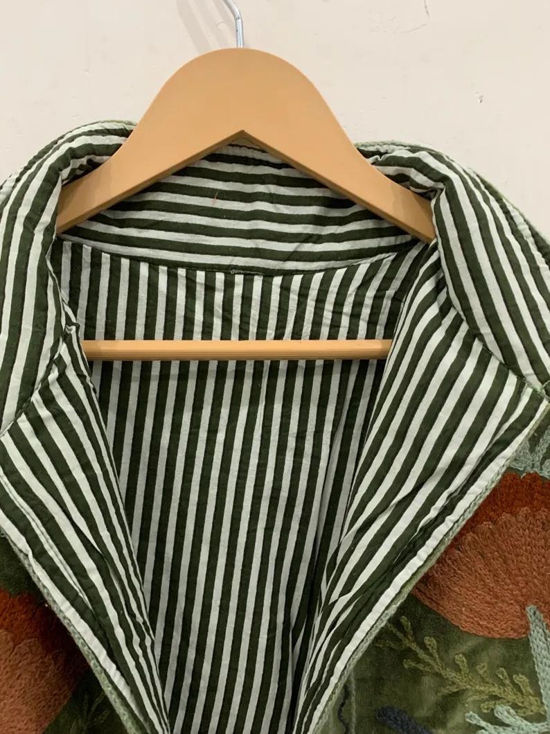 Pu&ograve; includere: Primo piano di una giacca verde con interno e colletto a righe. La giacca &egrave; appesa a una gruccia di legno. La giacca ha un motivo floreale ricamato nei toni dell'arancione e del verde.