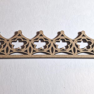 May include: A laser-cut wooden decorative trim piece featuring a repeating pattern of stylized crowns with floral and geometric cutouts. The trim is a light brown color and appears to be made of thin wood.