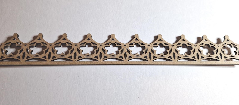 May include: A laser-cut wooden decorative trim piece featuring a repeating pattern of stylized crowns with floral and geometric cutouts. The trim is a light brown color and appears to be made of thin wood.