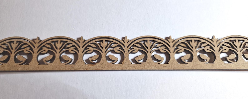 May include: A decorative wooden trim with a repeating pattern of arched designs, each featuring a stylized tree with leaves. The trim is a light brown color and has a delicate, openwork design, suitable for craft projects.