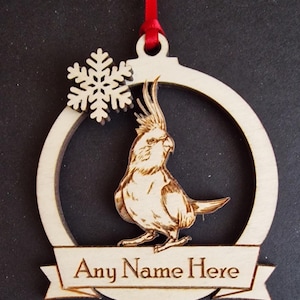 Puede incluir: Adorno navide&ntilde;o de madera con un grabado detallado de un ave cacat&uacute;a dentro de un marco circular. Tambi&eacute;n se incluye un copo de nieve y una pancarta con el texto "Any Name Here". El adorno cuelga de una cinta roja.