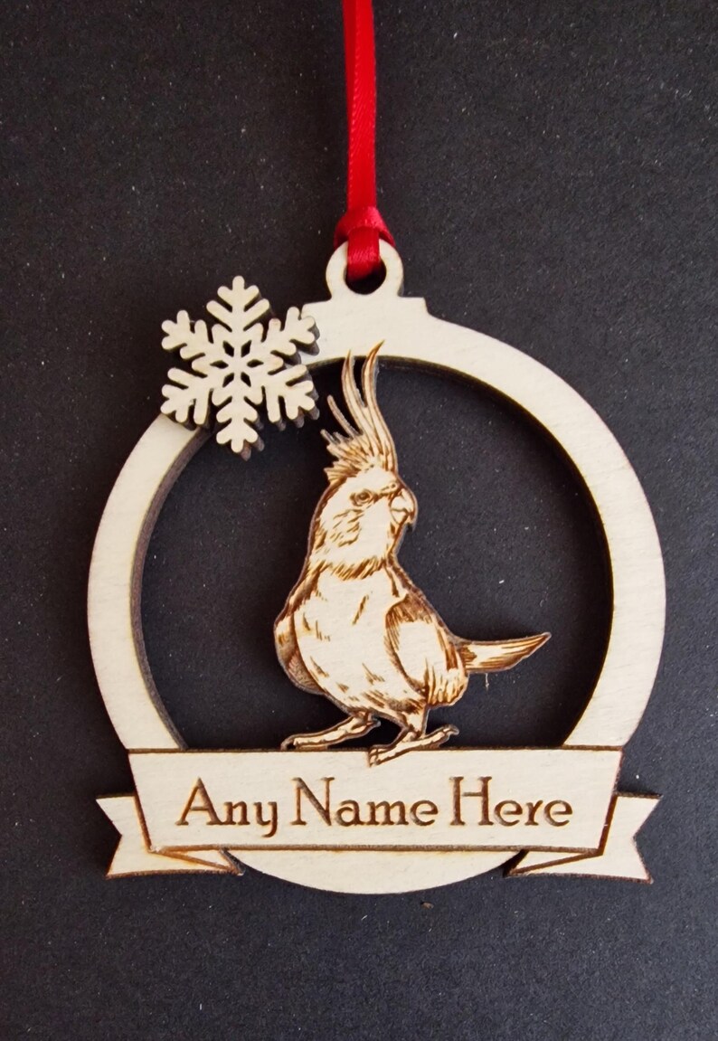 Puede incluir: Adorno navide&ntilde;o de madera con un grabado detallado de un ave cacat&uacute;a dentro de un marco circular. Tambi&eacute;n se incluye un copo de nieve y una pancarta con el texto "Any Name Here". El adorno cuelga de una cinta roja.