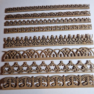May include: A collection of laser-cut wooden decorative borders in various ornate designs. The borders feature repeating patterns of arches, floral elements, and geometric shapes. The wood has a natural, light brown color, and the designs are intricately detailed.