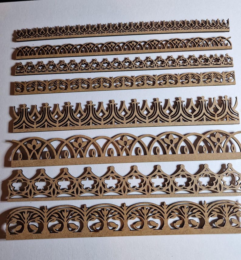May include: A collection of laser-cut wooden decorative borders in various ornate designs. The borders feature repeating patterns of arches, floral elements, and geometric shapes. The wood has a natural, light brown color, and the designs are intricately detailed.